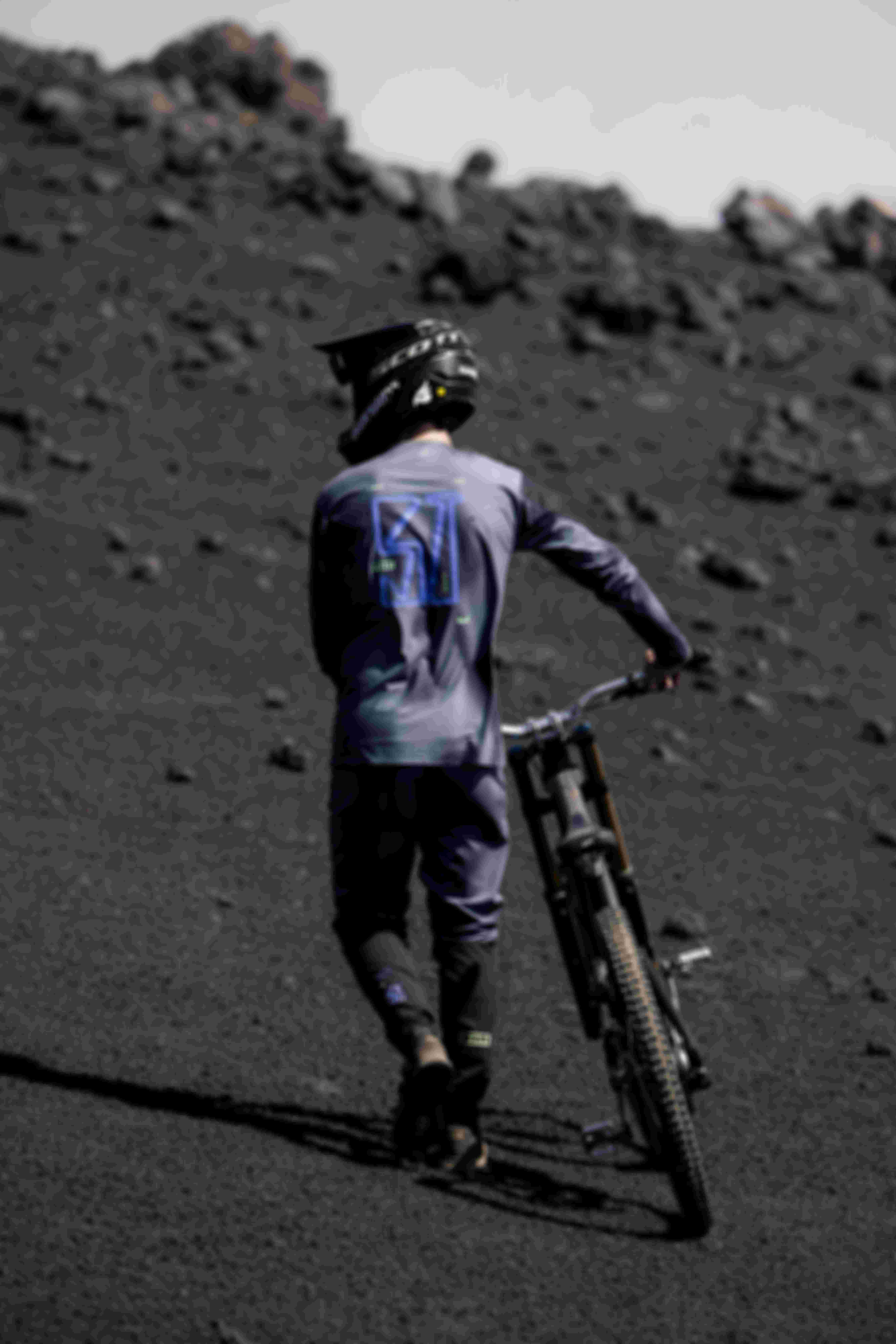 A person in cycling gear pushes a mountain bike up a rocky, barren slope, wearing a helmet and a long-sleeved shirt with the number 77 on the back.