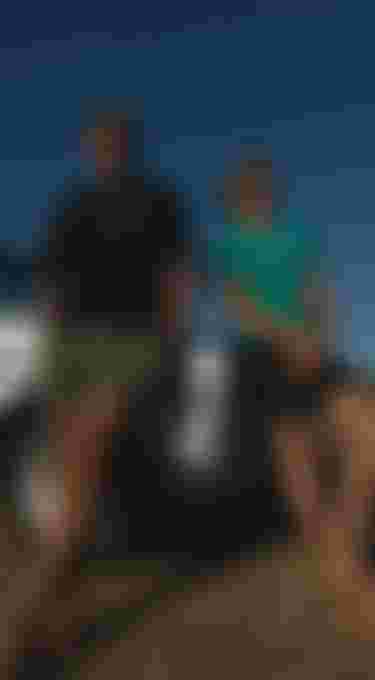 Close-up of a logo on black shorts; two people leaning against a white vintage car, standing on a sunny hill.