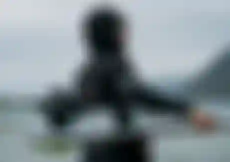 Woman in a hooded wetsuits carrying a kite surfboard on a misty beach