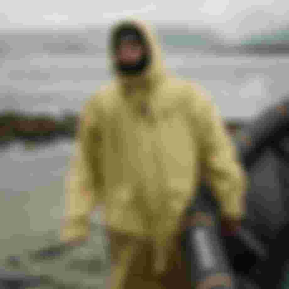 Person wearing a yellow rain coat with hood on the beach carrying a kite in one hand