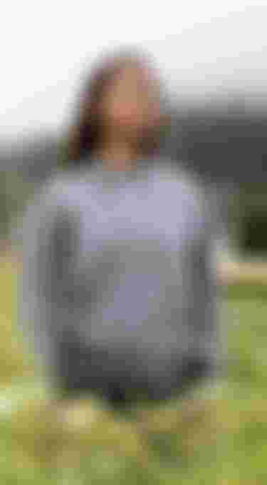 Woman wearing a grey sweater with ION logo standing in a grass field with a cloudy sky above