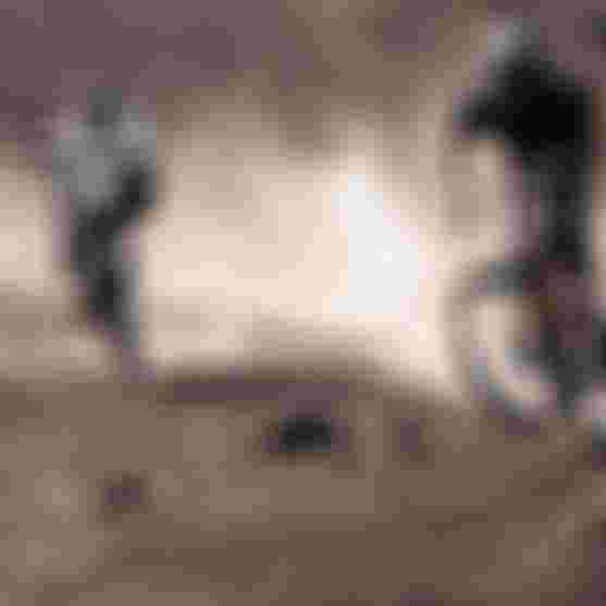 Two mountain bikers in helmets ride over rugged, dusty terrain, with one lifting the front wheel in an action-packed scene.