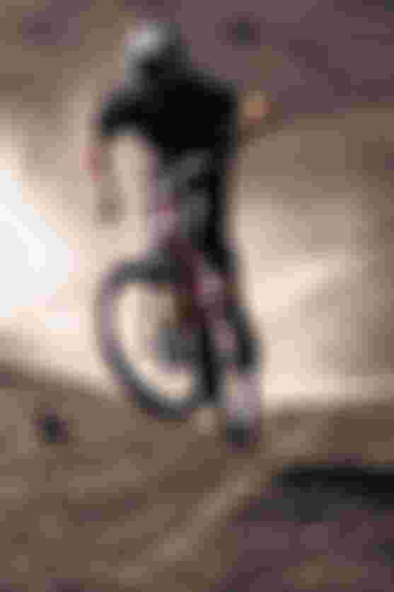 Two mountain bikers in helmets ride over rugged, dusty terrain, with one lifting the front wheel in an action-packed scene.