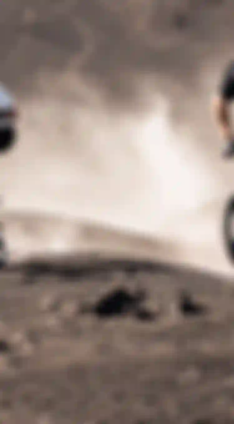 Two mountain bikers in helmets ride over rugged, dusty terrain, with one lifting the front wheel in an action-packed scene.