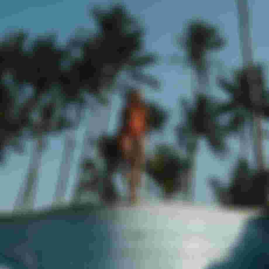 Woman in a red longsleeve shorty wetsuit standing at the edge of an empty pool with palm trees in the background