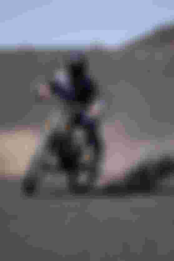 A mountain biker in protective gear rides on a dusty, barren landscape, kicking up dirt behind the bike under a clear blue sky.