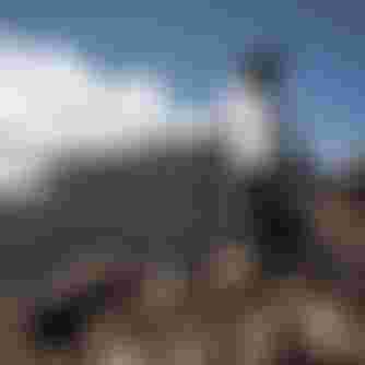 Mountain biker in gear stands with a bike on rocky terrain, overlooking a rugged landscape with a distant smoking volcano under a clear blue sky.