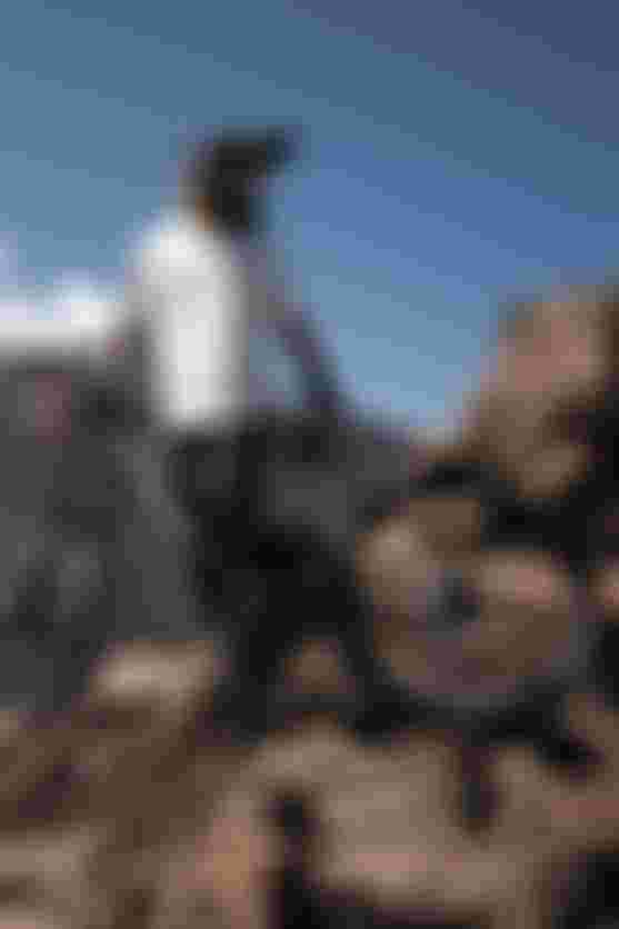 Mountain biker in gear stands with a bike on rocky terrain, overlooking a rugged landscape with a distant smoking volcano under a clear blue sky.