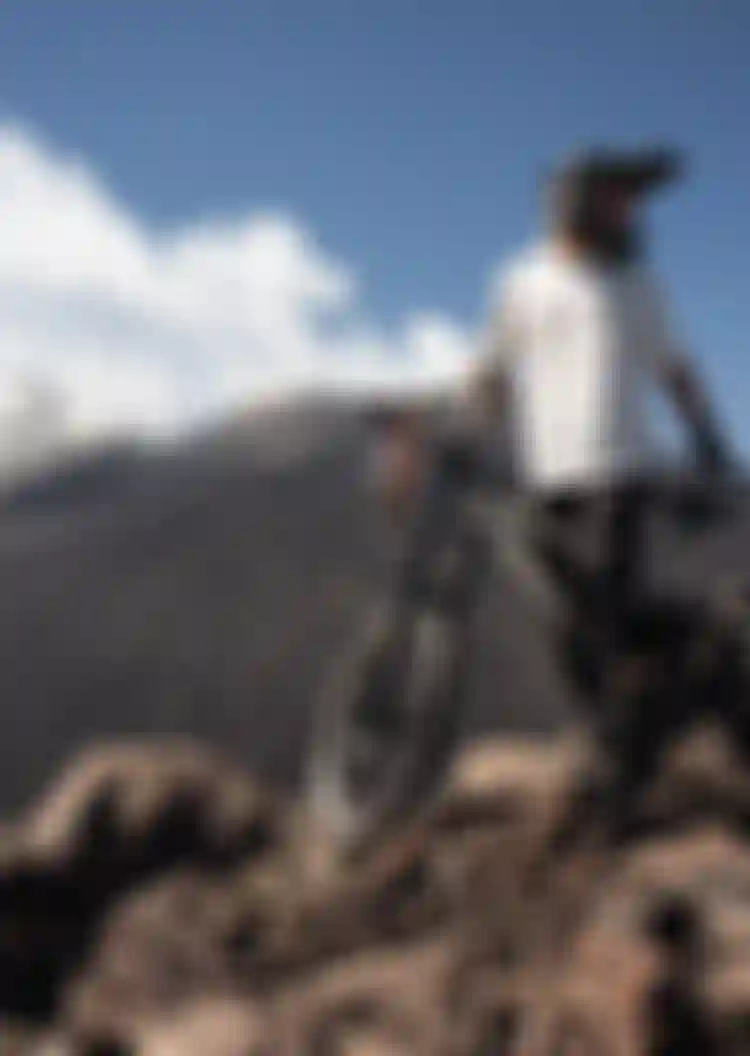 Mountain biker in gear stands with a bike on rocky terrain, overlooking a rugged landscape with a distant smoking volcano under a clear blue sky.