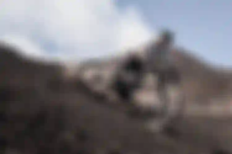 Cyclist in helmet rides a mountain bike down a steep, rocky slope, kicking up debris against a backdrop of hills and cloudy sky.