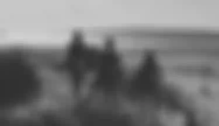 Three people in wetsuits carrying kite equipment walk toward a sandy beach by the ocean under a clear sky.