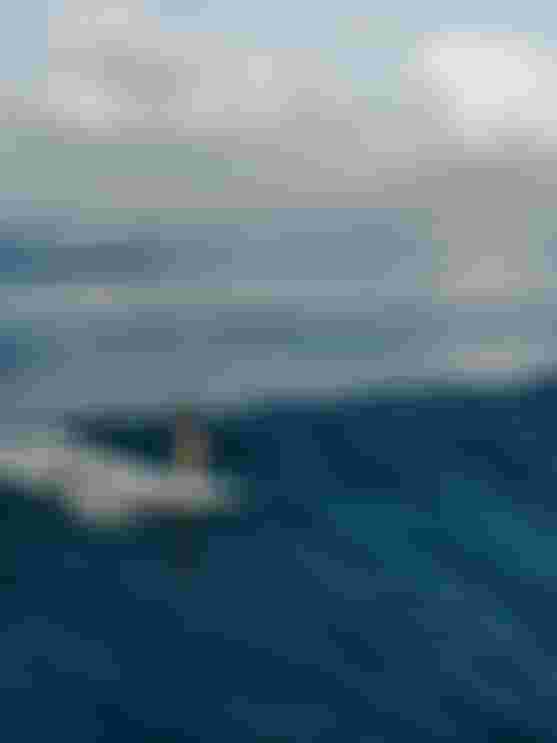 Surfer riding a wave on a calm ocean, blue sky with scattered clouds in the background.