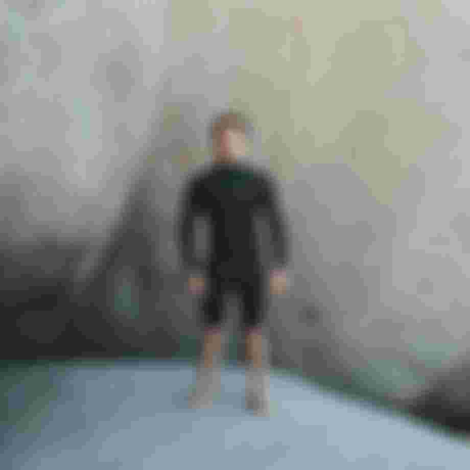 Man wearing a black wetsuit with short legs standing in front of a black rock looking directly into the camera
