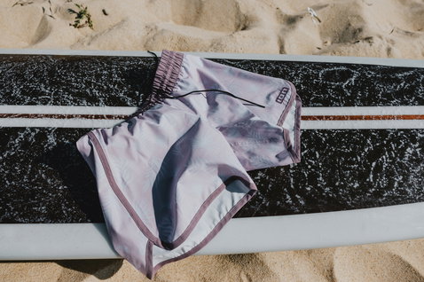 Boardshorts ION Mandiri donna in lavanda chiaro con bordo rosa su tavola da surf in spiaggia