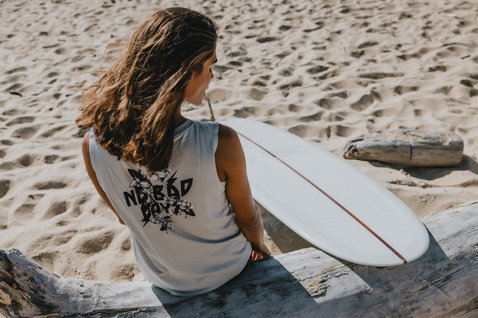 ION No Bad Days women's tank top in light grey relaxing at the beach with a surfboard
