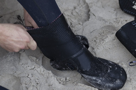 ION Ballistic Boots 3/2 neoprene boots in dark charcoal being put on at the sandy beach