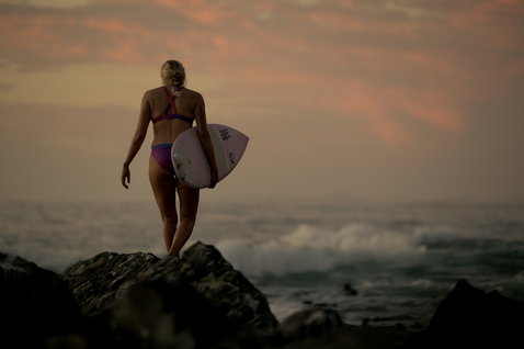 ION Surfkini women's bikini in magenta and purple during wave riding at rocky shoreline at golden hour