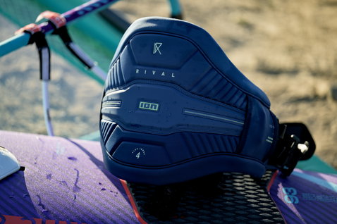 Arnês de assento ION Rival em azul meia-noite com cinto flex interno sobre prancha de windsurf na praia