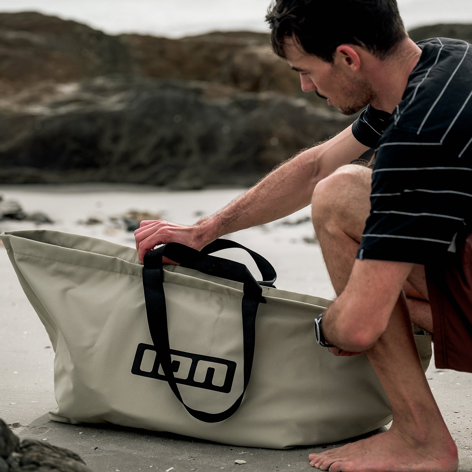 Sac de voyage ION Session Bag 80L en beige sable utilisé sur une plage rocheuse