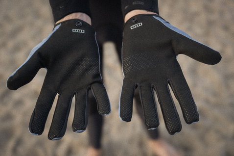 Gants doigts complets ION Amara en noir avec texture grip, portés aux deux mains sur la plage