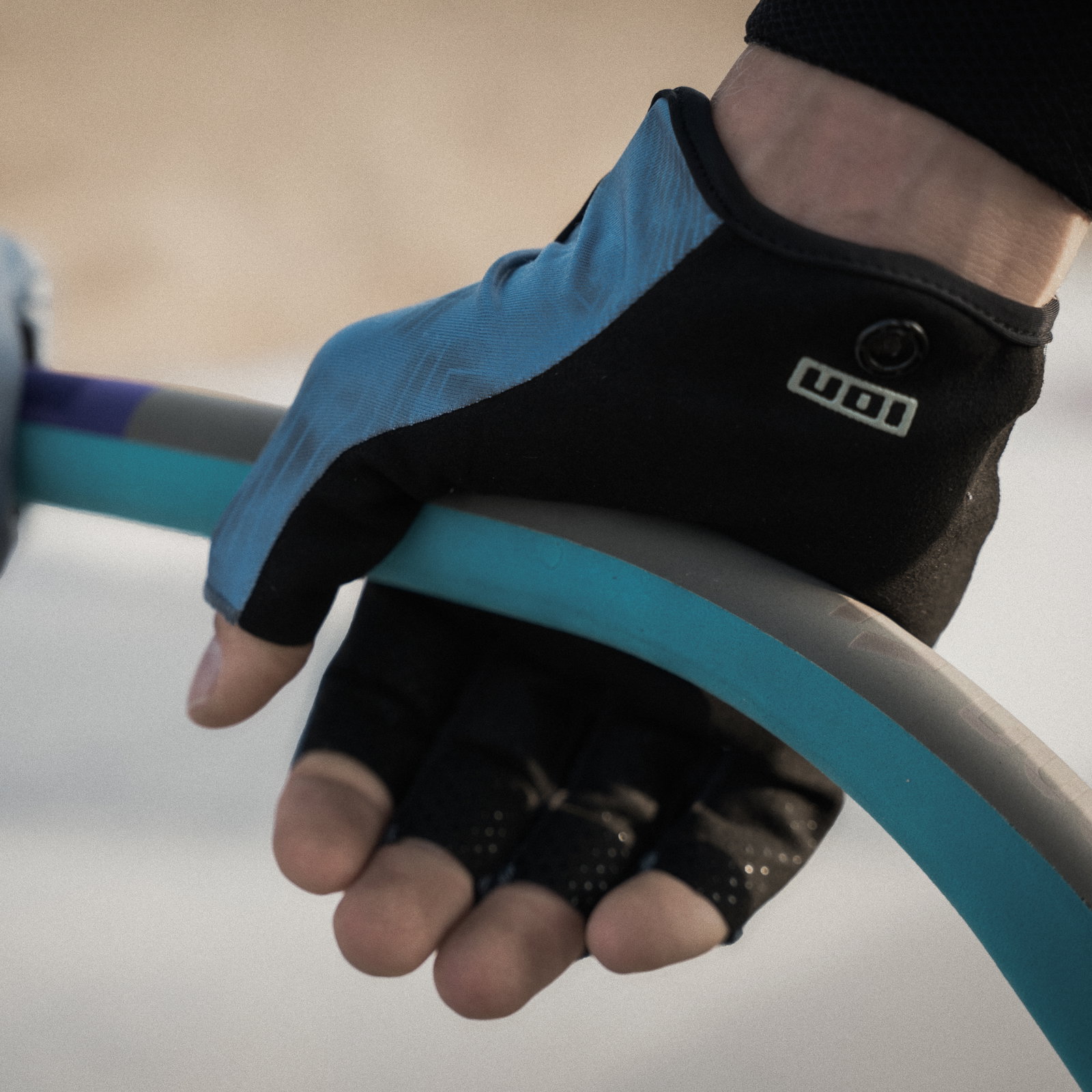 Gants ION Amara mi-doigt en noir et bleu acier saisissant un boom de windsurf