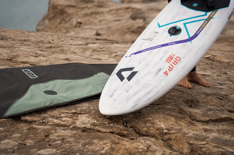 ION Core Stubby windsurf board bag on rocky coastline next to a white Duotone windsurf board