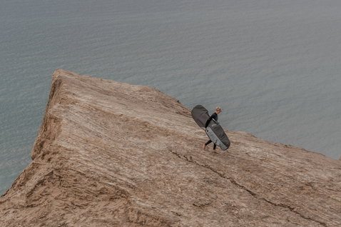 ION Core Stubby windsurf boardbag in charcoal grey carried along a coastal clifftop
