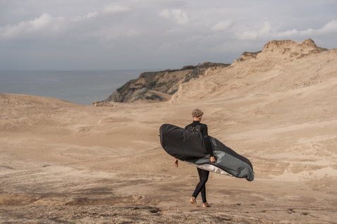 ION Core Stubby windsurf board bag carried over shoulder across coastal sand dunes