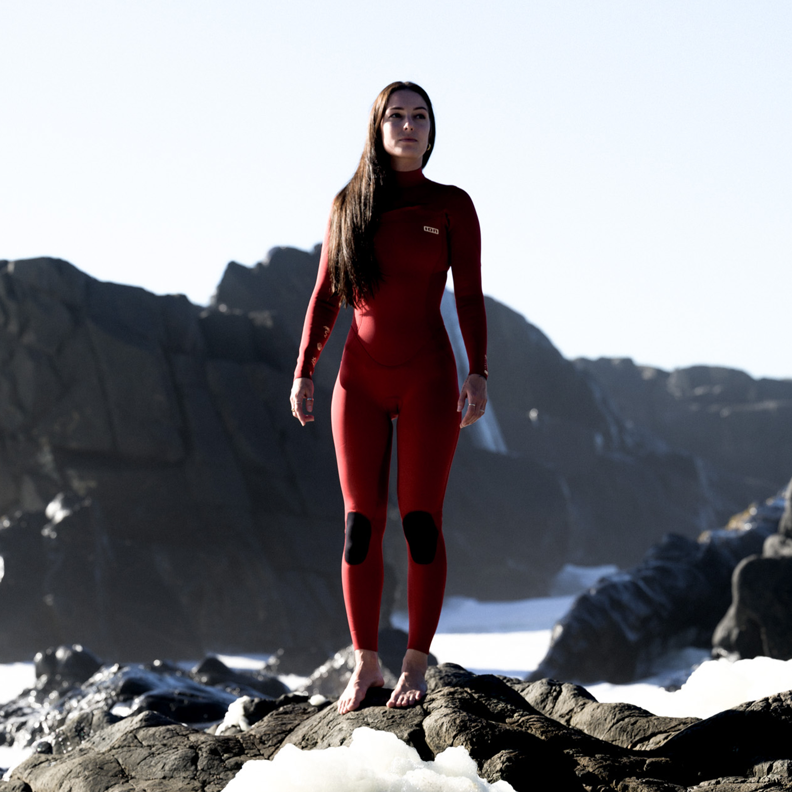 ION Amaze Core 3/2mm front-zip fullsuit in dark red worn by female model on coastal rocks