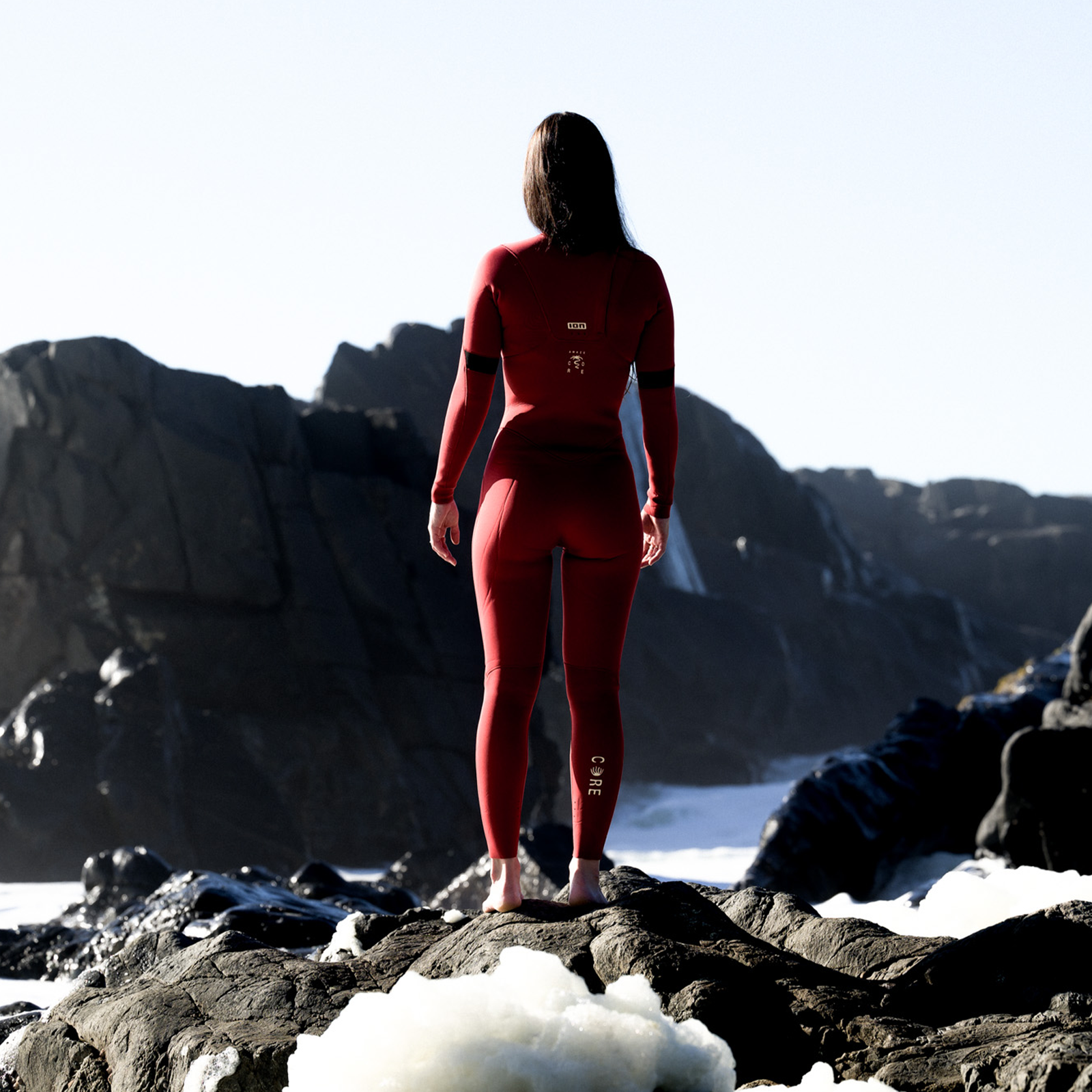 ION Amaze Core 4/3mm front-zip women's fullsuit in red during kitesurfing at a rocky coast
