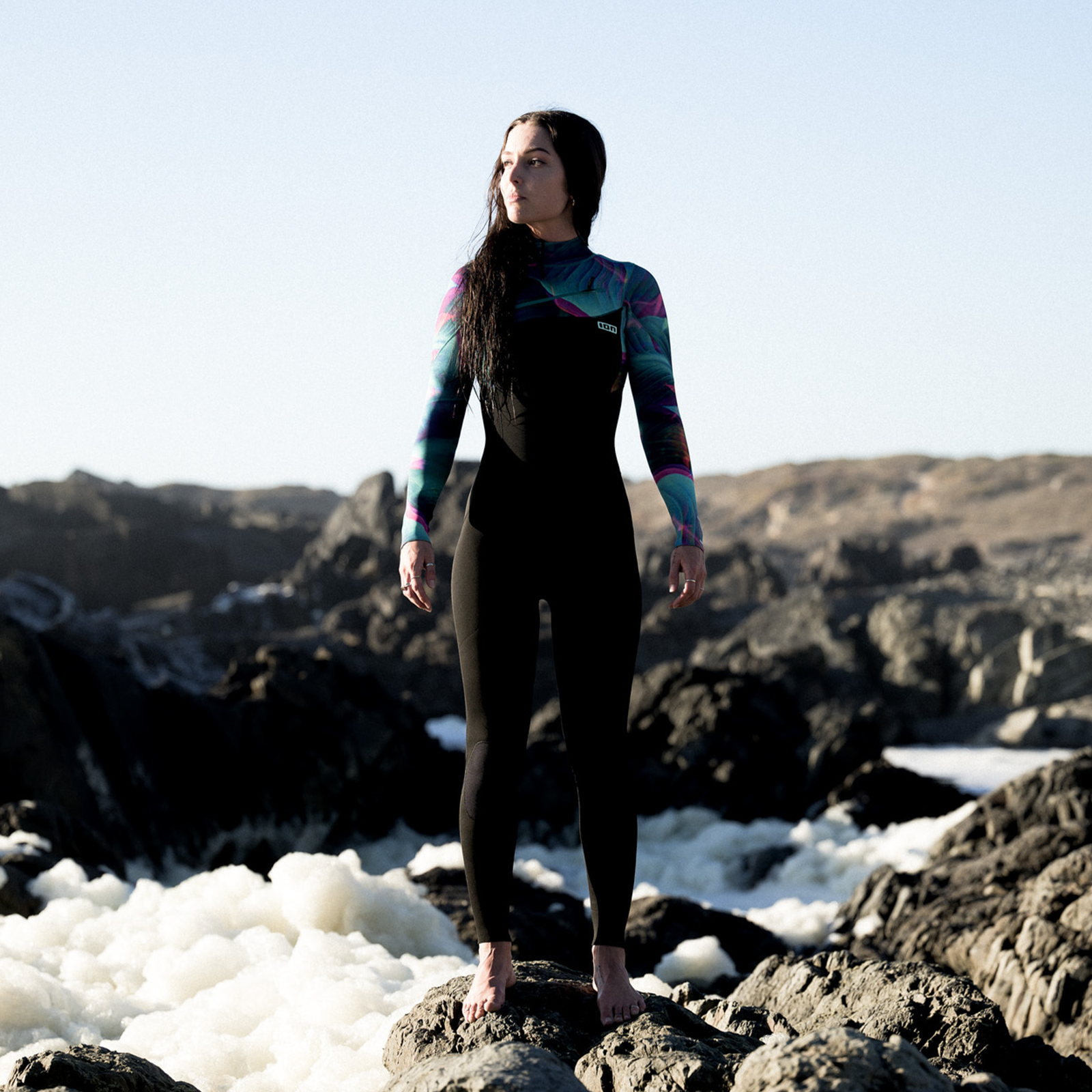 Muta ION Amaze Core 5/4 zip frontale in nero, turchese e rosa durante il surf su costa rocciosa