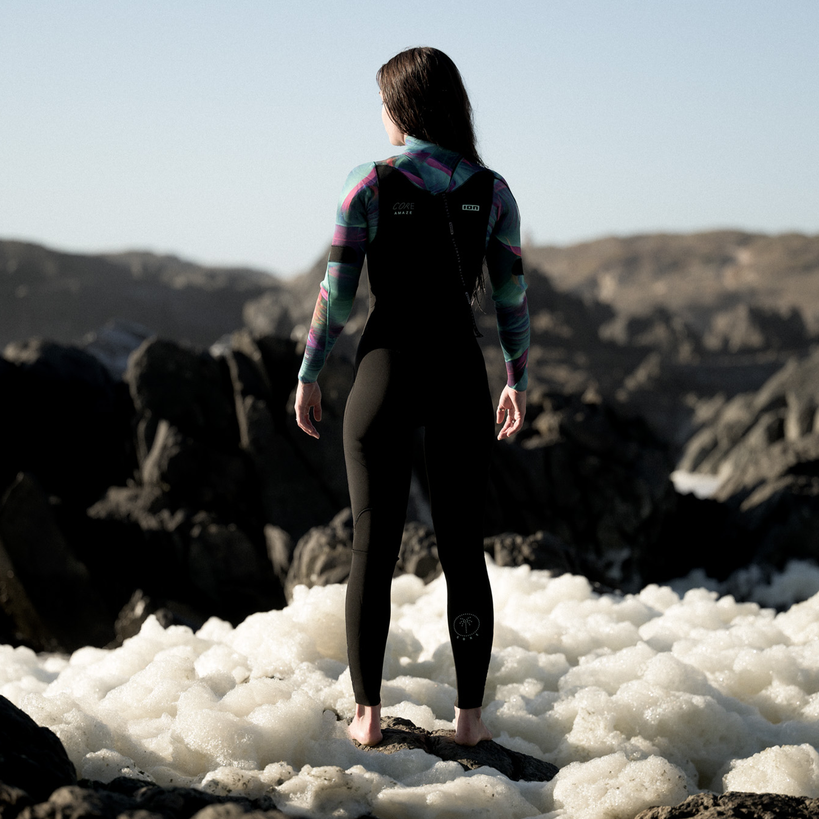 ION Amaze Core 5/4mm back-zip women's fullsuit wetsuit in black with teal and pink graphic, worn at rocky coastline