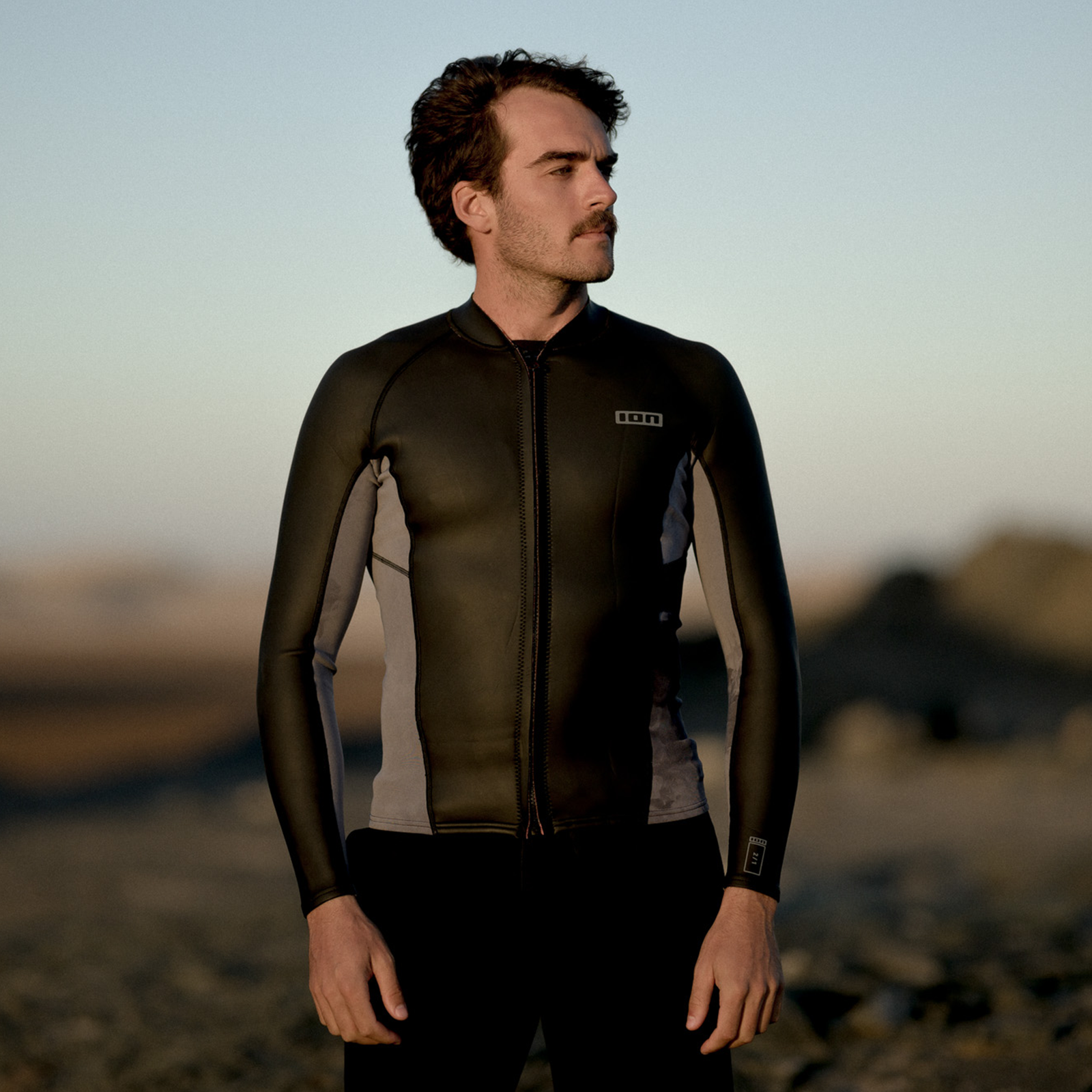 ION Neo Zip Top 2/1 LS Skin men wetsuit top in black olive and grey worn at the coast