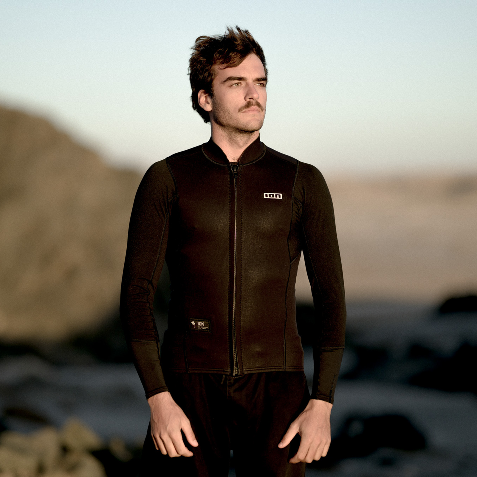 ION Neo Zip Top 2/1 LS men's neoprene top in dark brown-black at a rocky coastal beach