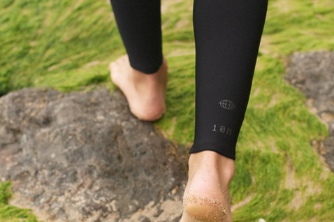 ION Capture Amp 6/5mm front-zip junior fullsuit in black walking barefoot on coastal rocks