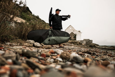 ION Wing Boardbag Core Slim in black and olive green carried on a rocky coastal beach