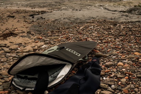 ION Wing Boardbag Core Slim open on a rocky pebble beach with ocean waves in the background