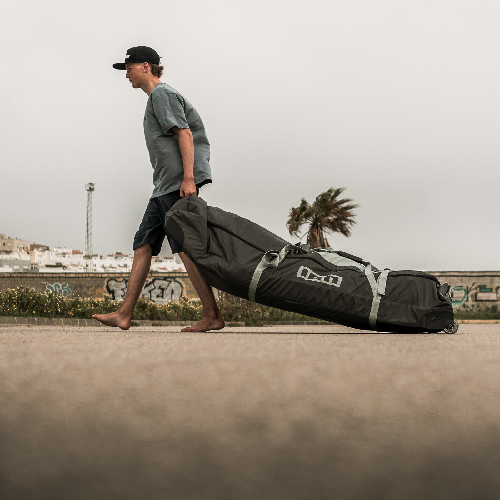 ION Gearbag Core wheeled kite gear bag in dark charcoal being pulled along a coastal promenade