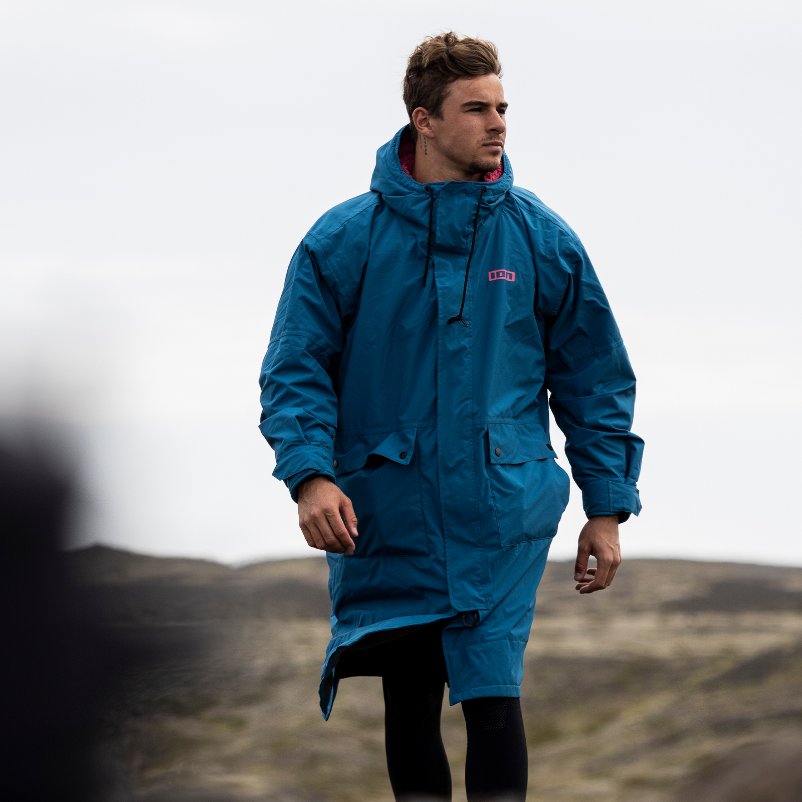 ION Storm Coat water jacket in ocean blue worn by a model on a rocky coastal hillside