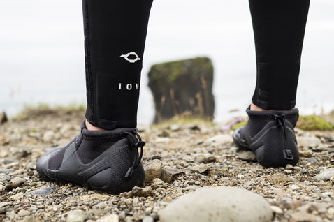 ION Plasma Shoes 2.5 Round Toe neoprene boots in black during kitesurfing at a rocky coastline