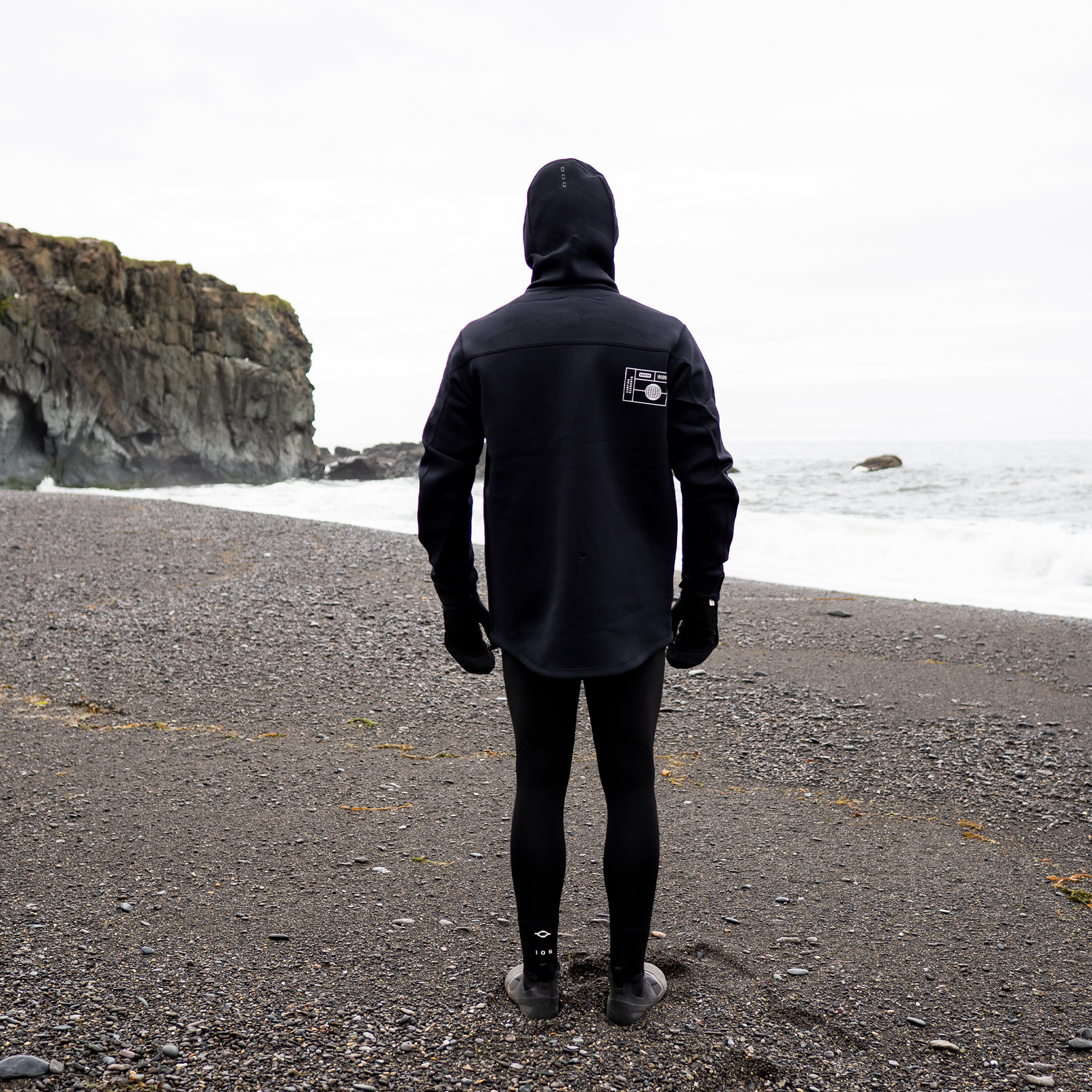 ION Neo Shelter Jacket Core veste en néoprène à capuche en noir sur une côte rocheuse