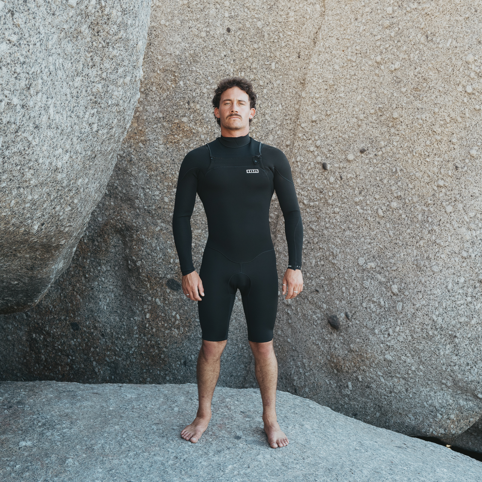 ION Element 2/2mm front-zip long-sleeve shorty wetsuit in black worn at rocky coastal boulders