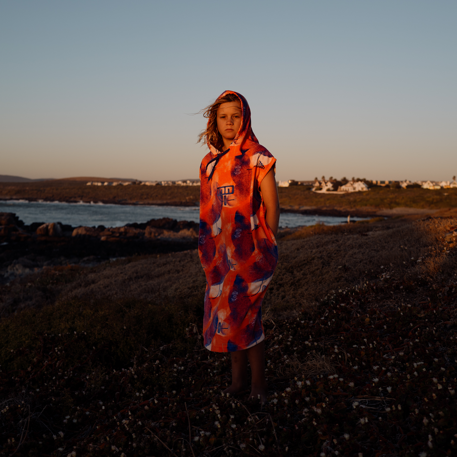 ION Poncho Grom kids surf poncho in orange-red tiger print at the rocky coastline