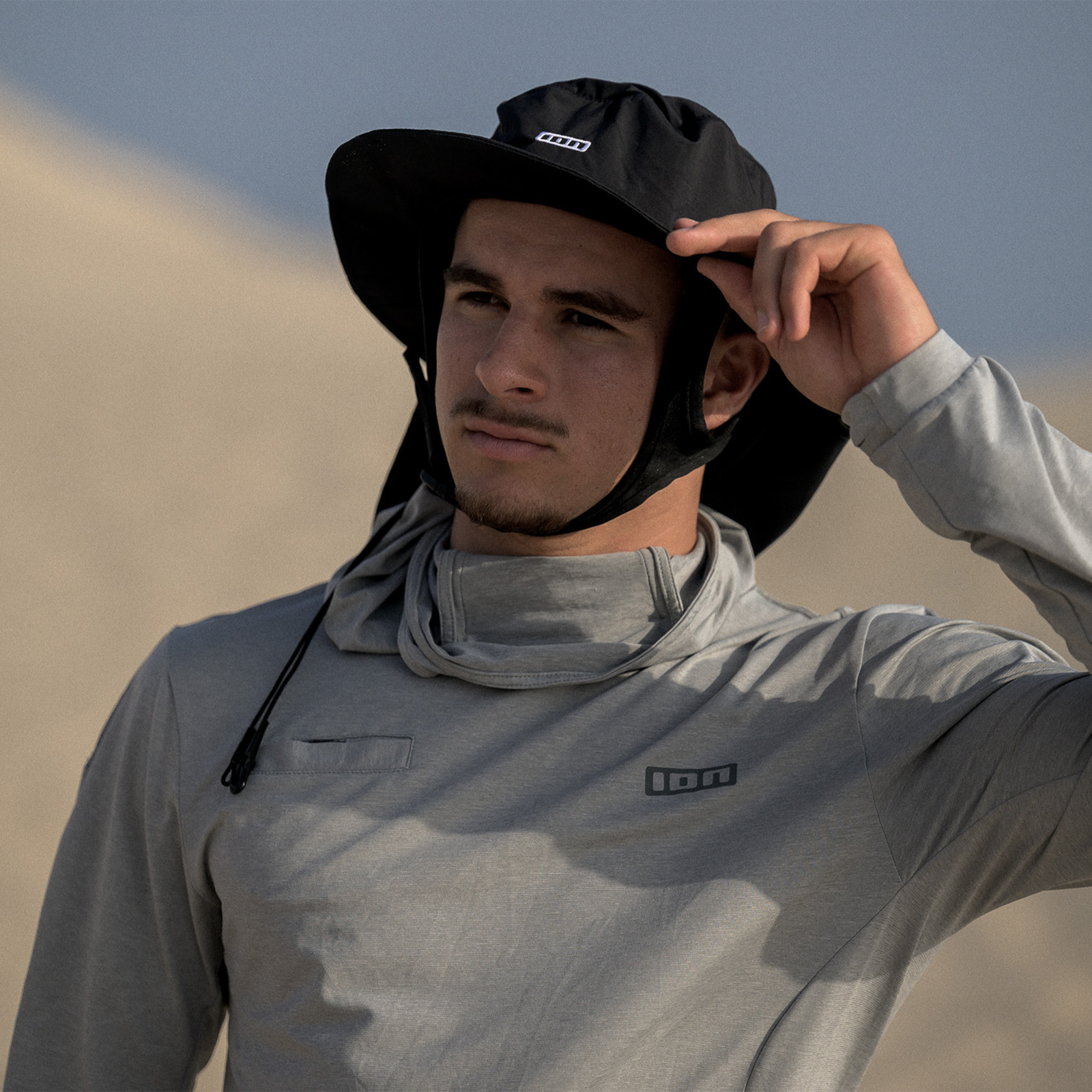 ION Water Cap Beach Hat in black with wide brim and chin cord, worn by a man in front of sandy dunes