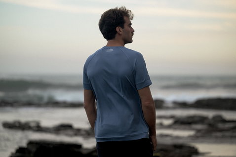 ION Wetshirt short sleeve men in slate blue during kitesurfing at a rocky coastline