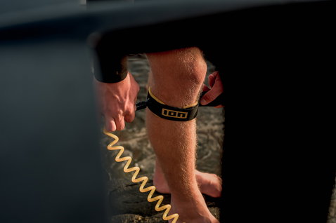 ION Wing Board Leash Core Coiled Knee in nero e oro durante il wing foiling sull'acqua