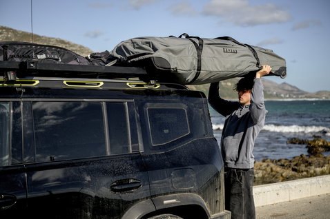 ION Wing Gearbag Core travel bag in grey being loaded onto a car roof rack at the coast