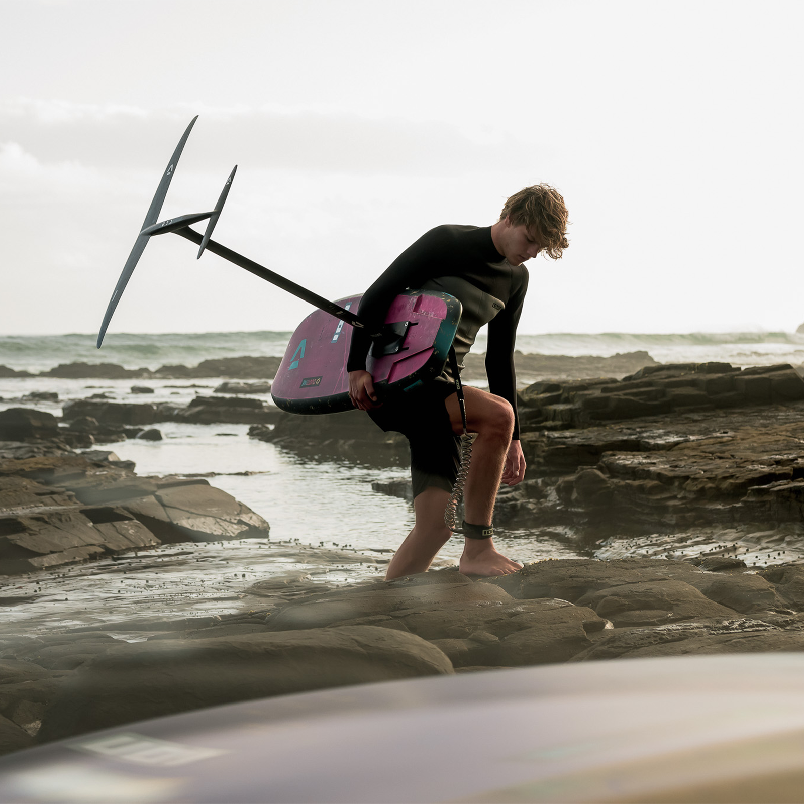 ION Wing Board Leash Core coiled ankle leash during wing foiling at a rocky coastal shore