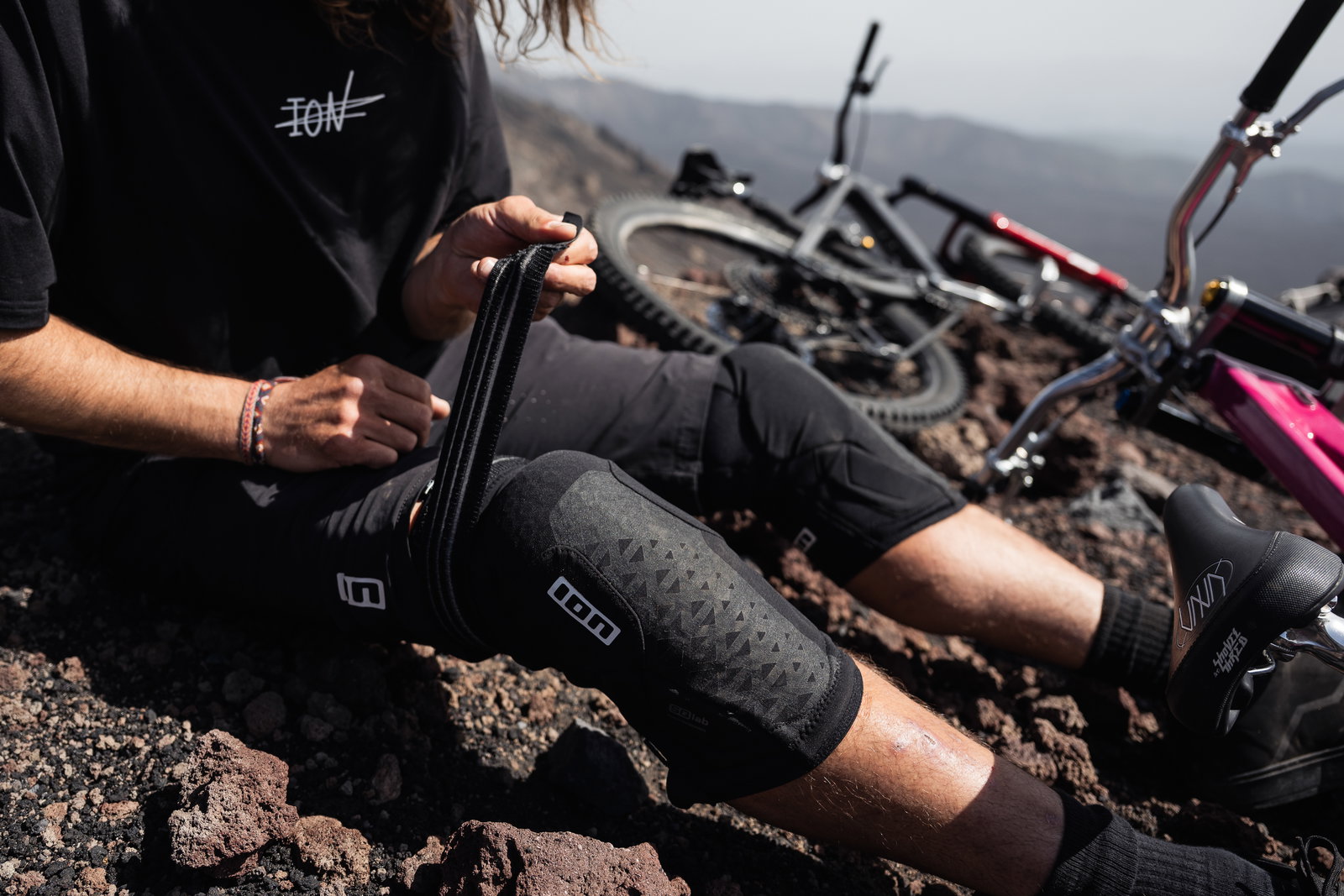 ION K-Pact knee pads in black being fitted on rocky volcanic terrain during mountain biking