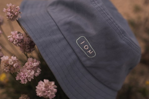 ION Bucket Hat em azul ardósia entre flores silvestres rosas ao ar livre